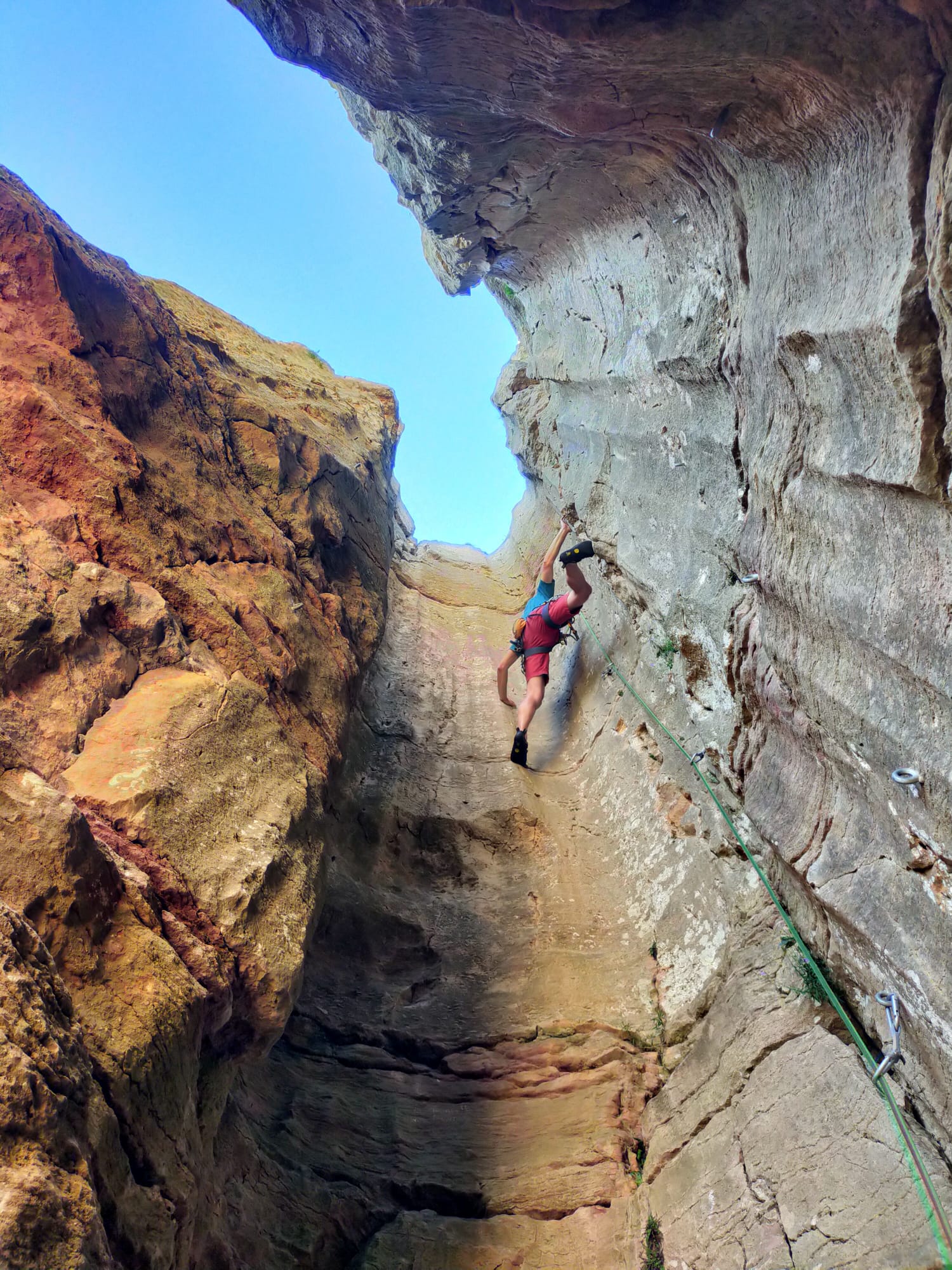 Mikaël, encordé, progresse en dévers dans un couloir rocheux étroit, entre falaise ocre et paroi grise, sous une arche de pierre